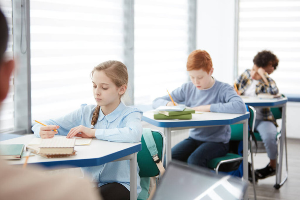 Zijaanzicht bij multi-etnische groep kinderen die in de klas achter elkaar achter een bureau zitten en schrijven of toetsen doen, kopieerruimte - Foto, afbeelding