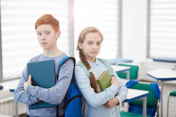 Vyötärö ylös muotokuva punatukkainen teini poika ja tyttö seisoo selkä selkää vasten, kun poseeraa koulussa luokkahuoneessa ja pitää kirjoja, kopioi tilaa - Valokuva, kuva