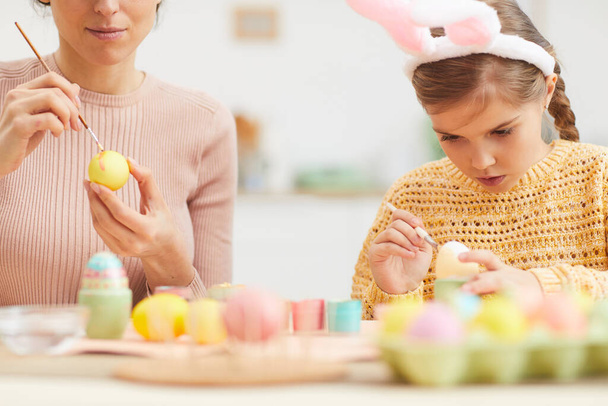 Portrait of cute little girl painting Easter eggs with mother while wearing bunny ears in cozy kitchen interior, copy space - Photo, Image