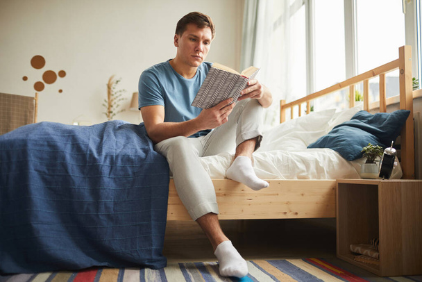 Meleg tónusú teljes hosszúságú portré modern fiatalemberről, aki könyvet olvas, miközben otthon ül az ágyon, másolja a helyet - Fotó, kép