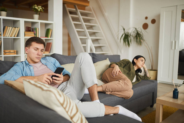Retrato de larga duración de la pareja joven aburrida viendo la televisión en el sofá en casa, se centran en el hombre usando el teléfono inteligente en primer plano, espacio de copia
 - Foto, Imagen