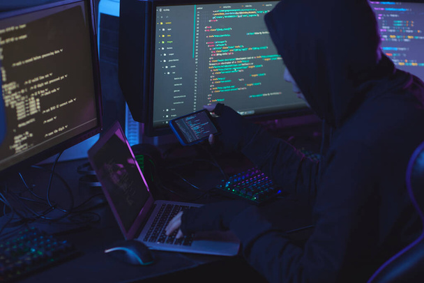 Vista trasera en el hacker de seguridad cibernética irreconocible con capucha mientras se trabaja en la programación en la habitación oscura, espacio de copia
 - Foto, Imagen