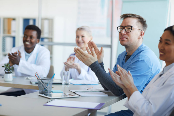 Grupo multiétnico de médicos aplaudiendo mientras están sentados en la mesa durante la conferencia médica, se centran en el hombre maduro en primer plano, espacio para copiar
 - Foto, Imagen
