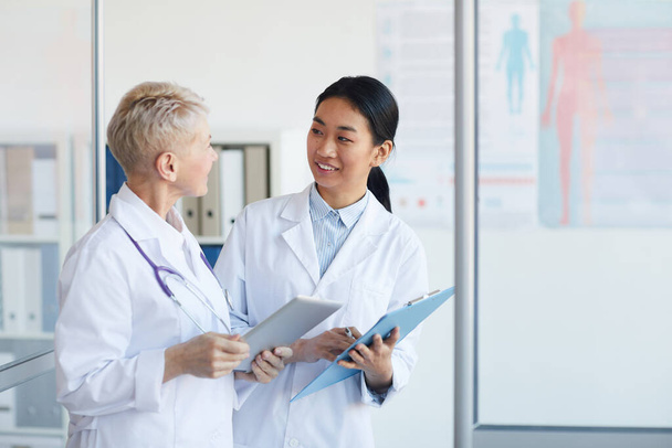 Waist up portrait of young female doctor talking to supervisor and smiling cheerfully while standing in medical office interior, copy space - Photo, Image