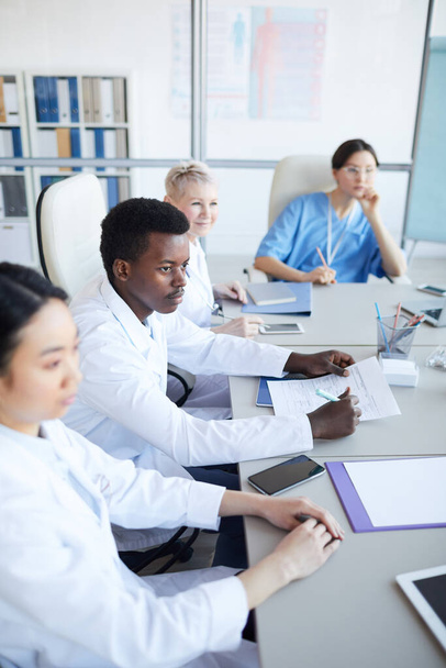 Высокий угол обзора у молодого афроамериканского врача, сидящего за столом во время медицинской конференции с коллегами, копировальное пространство
 - Фото, изображение