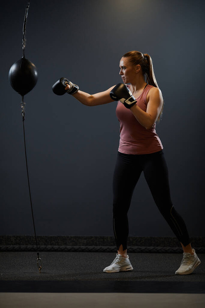 Teljes hosszúságú portré eltökélt fiatal nő boksz boxzsák ellen fekete háttér harc edzés vagy edzés során, másolás tér - Fotó, kép