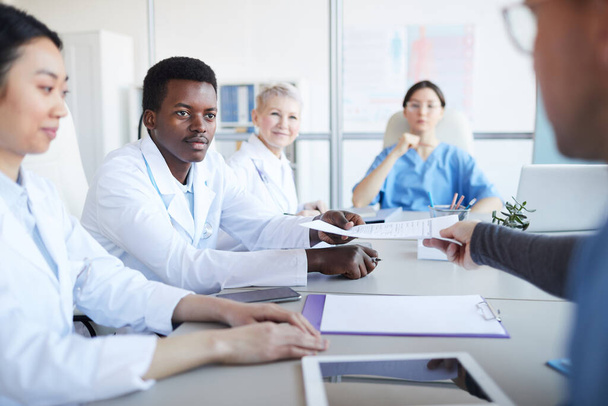Портрет молодого афроамериканского врача, заключившего договор или документ, сидящего за столом во время медицинской конференции с коллегами, копировальное пространство
 - Фото, изображение