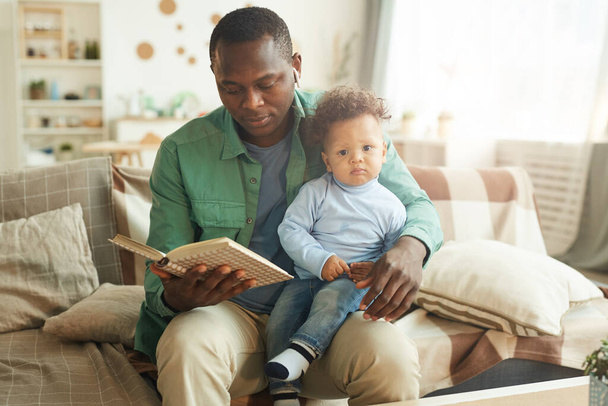Портрет взрослого афроамериканца, читающего книгу ребенку, сидящему на коленях отцов, копирующего пространство
 - Фото, изображение