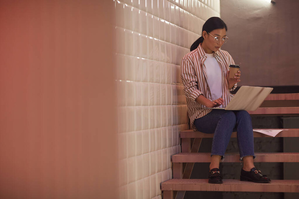 Pełna długość portret nowoczesnej Azjatki za pomocą laptopa podczas siedzenia na klatce schodowej przez kafelki ściany, przestrzeń kopiowania - Zdjęcie, obraz