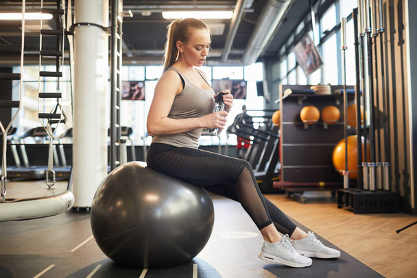 Retrato de comprimento total da jovem esportiva sentada na bola de fitness preta no interior ginásio moderno, espaço de cópia
 - Foto, Imagem