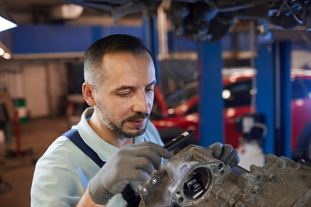 Retrato de ángulo alto del mecánico del coche barbudo que inspecciona la caja de cambios en el taller de reparación de automóviles, espacio de copia
 - Foto, Imagen
