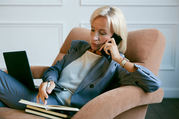 Portrait of modern mature businesswoman working with laptop and speaking by phone while siting in chair in office, copy space - Valokuva, kuva