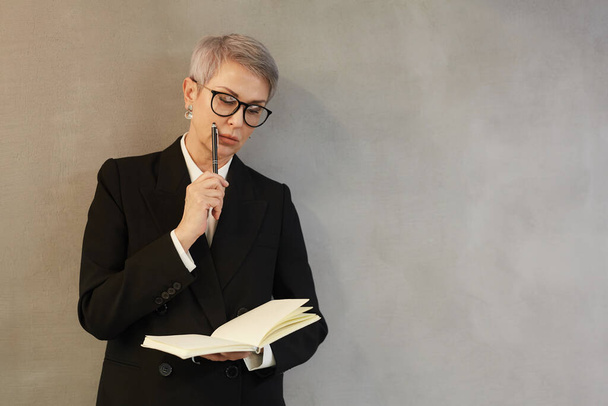 Minimal waist up portrait of successful mature businesswoman writing notes in planner while standing against gray wall and wearing black suit, copy space - Foto, Imagem