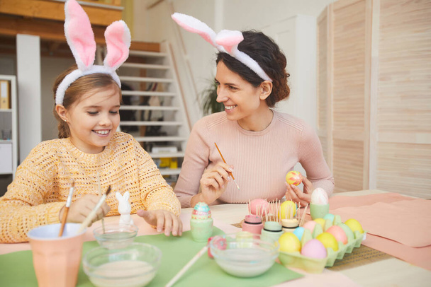 Portrait of happy young mother and daughter painting Easter eggs in cozy kitchen interior, both wearing bunny ears, copy space - 写真・画像