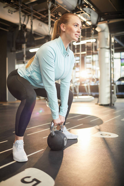 Full length portrait of smiling young woman picking up dumbbell during strength workout in sunlit gym, copy space - Photo, Image