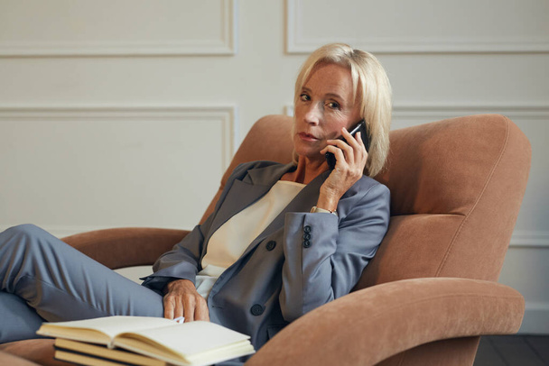 Retrato mínimo de la exitosa mujer de negocios madura mirando a la cámara mientras habla por teléfono en el sillón en la oficina, espacio de copia
 - Foto, Imagen