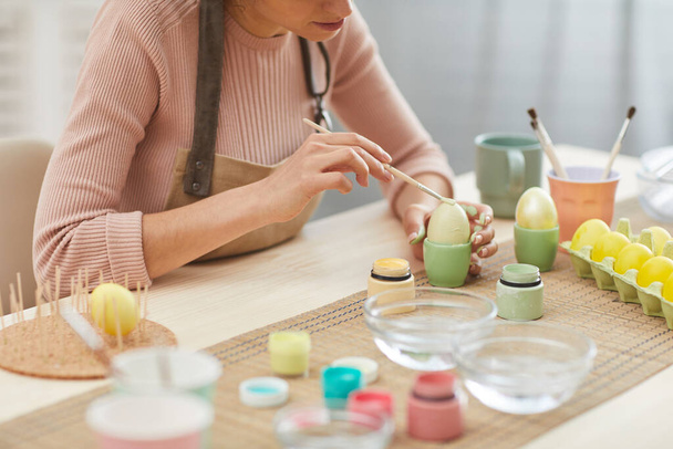 Cropped portrait of unrecognizable young woman painting eggs in pastel colors for Easter while sitting at table in kitchen or art studio, copy space - Photo, Image