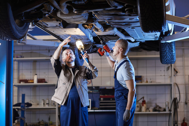 Portrait de deux mécaniciens matures debout sous la voiture sur ascenseur tout en inspectant le véhicule dans l'atelier de réparation automobile, espace de copie
 - Photo, image