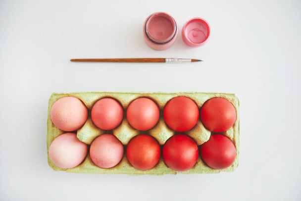 Κάτοψη παστέλ ροζ και κόκκινων πασχαλινών αυγών σε καφάσι τοποθετημένα σε μίνιμαλ σύνθεση με πινέλο σε λευκό φόντο, αντίγραφο χώρου - Φωτογραφία, εικόνα