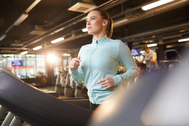 Retrato de uma jovem esportiva correndo em esteira rolante durante o treino cardio no ginásio moderno, brilho da lente
 - Foto, Imagem