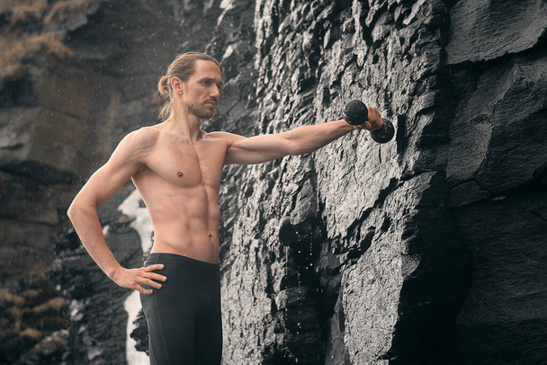 Sporty man doing exercises with dumbbells. Photo of muscular model with naked torso on rocky background. Fitness and healthy lifestyle concept - Φωτογραφία, εικόνα