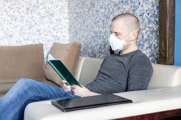Młody człowiek z respiratorem ffp1 na temat samoizolacji w swoim mieszkaniu podczas kwarantanny koronawirusu covid-19 czyta książkę - Zdjęcie, obraz