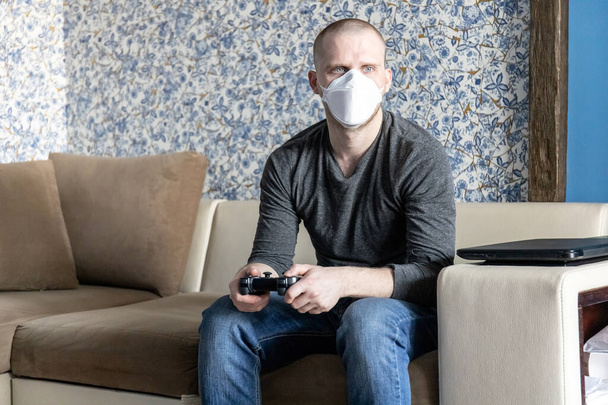 nuori mies, jolla on ffp1-hengityssuojain karanteenissa asunnossaan coronavirus covid-19 pandemian aikana, pelaa pelikonsolia
 - Valokuva, kuva