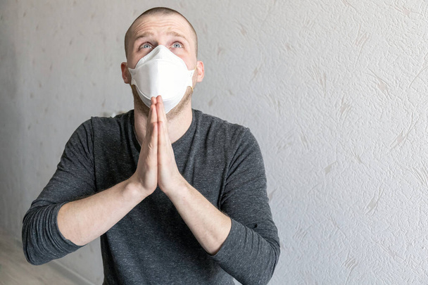 Młody człowiek z respiratorem ffp1 w kwarantannie w swoim mieszkaniu podczas pandemii koronawirusa covid-19 modli się z zaciśniętymi dłońmi - Zdjęcie, obraz