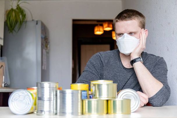 young man in medical mask at home with canned food, preparing for a pandemic, home quarantine - Photo, Image