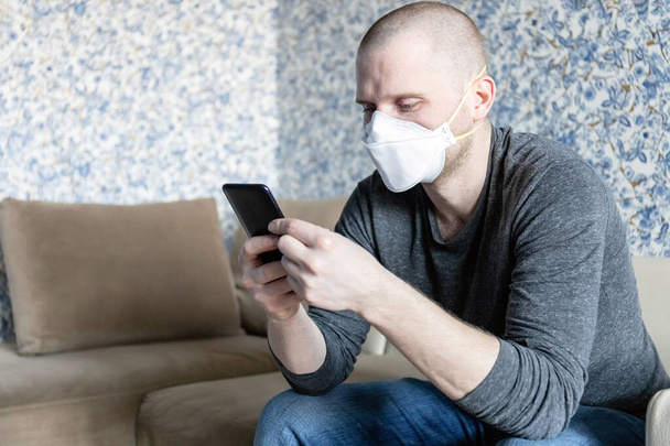 νεαρός άνδρας με αναπνευστήρα ffp1 σε αυτο-απομόνωση στο διαμέρισμά του κατά τη διάρκεια καραντίνας του coronavirus covid-19 διαβάζει ειδήσεις σε ένα smartphone - Φωτογραφία, εικόνα
