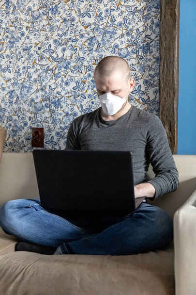 Młody człowiek z respiratorem ffp1 w kwarantannie w swoim mieszkaniu podczas pandemii koronawirusowej, Covid-19 pracujący na laptopie siedząc na kanapie - Zdjęcie, obraz