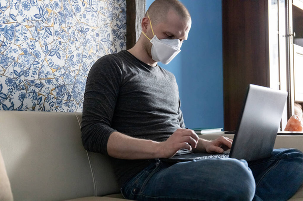 young man with a ffp1 respirator in quarantine in his apartment during a coronavirus pandemic, Covid-19 working on a laptop while sitting on a sofa - Photo, Image
