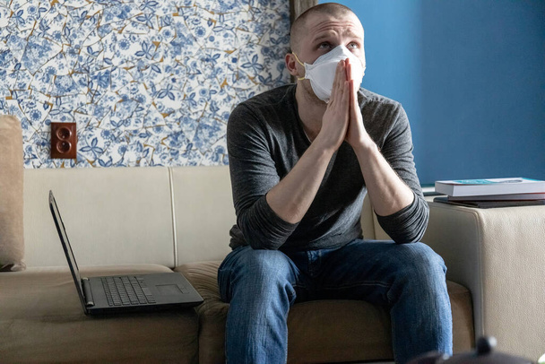 Молодой человек с респиратором ffp1 в карантине в своей квартире во время пандемии коронавируса ковид-19 молится сжимая руки
 - Фото, изображение