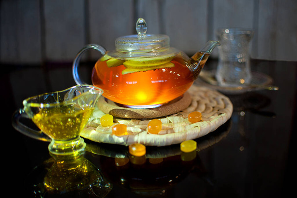 Чай с грейпфрутом и специями в прозрачном чайнике на деревянном подставке на темном фоне
 - Фото, изображение
