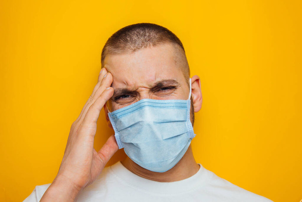 The guy holds on to his head, indicating that it hurts. Attractive man in a mask looks at the camera. Colds, flu, virus, tonsillitis, acute respiratory infections, quarantine, epidemic concept - Zdjęcie, obraz