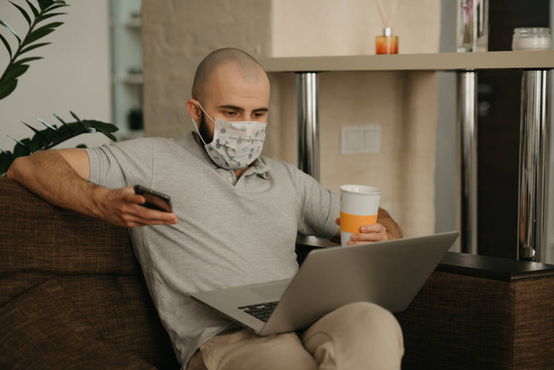 Un uomo con una maschera facciale che lavora a distanza sul portatile durante la quarantena per evitare il coronavirus diffuso. Un ragazzo lavora da casa con un telefono e una tazza di tè durante la pandemia del COVID-19
. - Foto, immagini