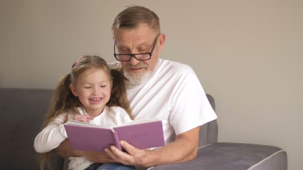 Grandfather teaches his granddaughter to read books. Family educational concept. An elderly man and a little girl leaf through a textbook - Filmagem, Vídeo
