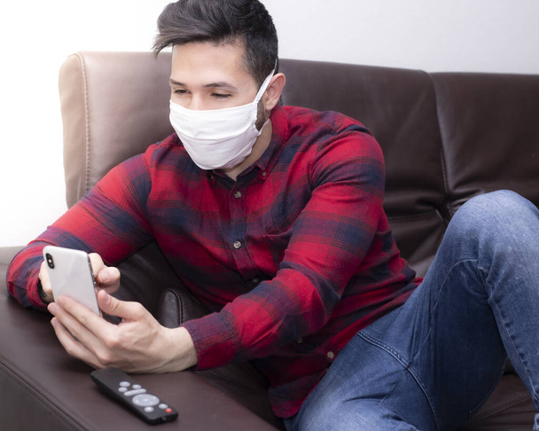 Young man with protective mask interacting with his smartphone indoors for quarantine. Self-insulation for Covid-19 - Foto, Imagen