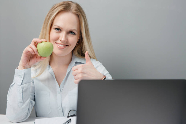 Kind-hearted girl smiling looks at the camera and holds an apple in her hand while sitting behind her workplace in the office. Copy space - Фото, зображення