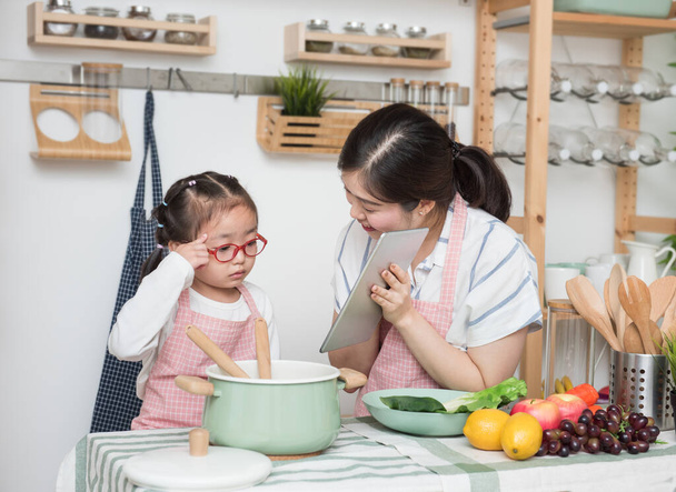 Asian woman mother and daughter play together in kitchen,mom hold tablet for teach little girl how to cook in semester break,social distancing, stay home concept. - Foto, Imagen