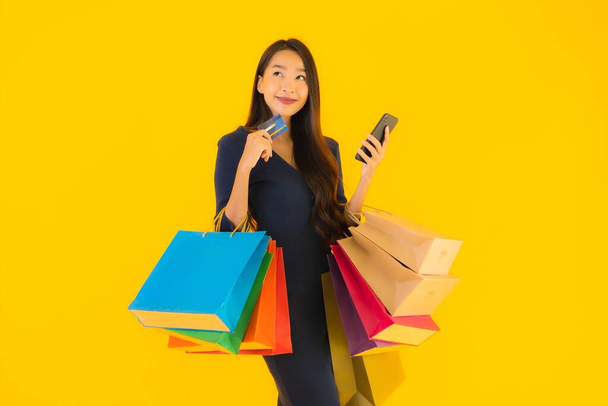 Portrét krásná mladá asijská žena s nákupní taškou kreditní karty a chytrý mobilní telefon žluté izolované pozadí - Fotografie, Obrázek