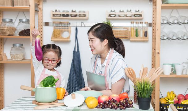 Азійська жінка, мати і дочка грають разом на кухні, мати тримає планшет, щоб навчити маленьку дівчинку готувати на перерві семестру, соціальну відстань, залишатись вдома концепт. - Фото, зображення