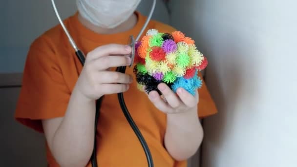 a boy in a mask, with brown hair, in an orange t-shirt sits on the stairs in the house and listens with a stethoscope to toys that look like a coronavirus with spines, a large bundle of thorns - Filmagem, Vídeo