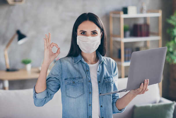 Photo of pretty lady stay home alone quarantine hold notebook wear protect anti virus mask showing okey symbol distance study work remote living room indoors - Foto, afbeelding