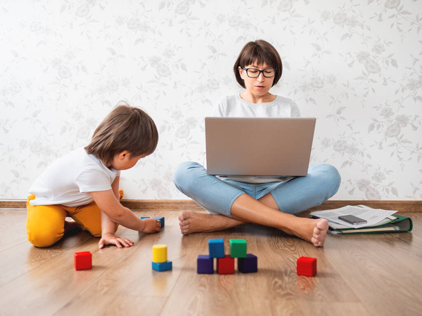 Mom and son argue at home quarantine because of coronavirus COVID19. Mother remote works with laptop, son plays with toy blocks. Self isolation at home. - Фото, изображение