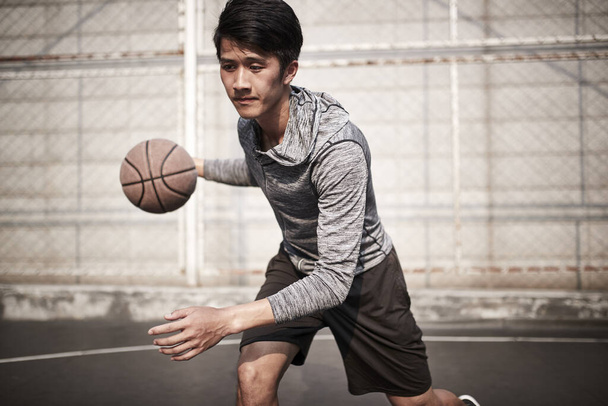 nuori aasialainen mies koripalloilija harjoittelee pallon käsittelytaitoja ulkona kentällä
 - Valokuva, kuva