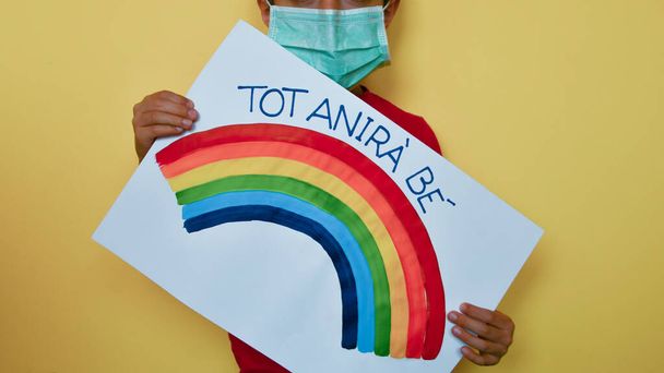 A young boy wears a disposable green mask with the caption todo ira bien and rainbow on a oblique sheet of paper in front of him - Photo, Image