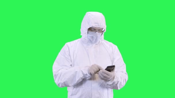 Masked man in protective suit surfing the phone while standing against green background - Video