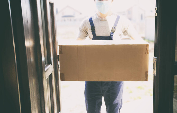 The courier is delivering the carton box to the home  wearing latex gloves and medical mask - Фото, изображение