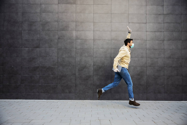 Un hombre casual corriendo contra una pared negra saludando. Lleva una máscara protectora y una tableta. Prevención de la enfermedad epidémica por coronavirus
. - Foto, Imagen